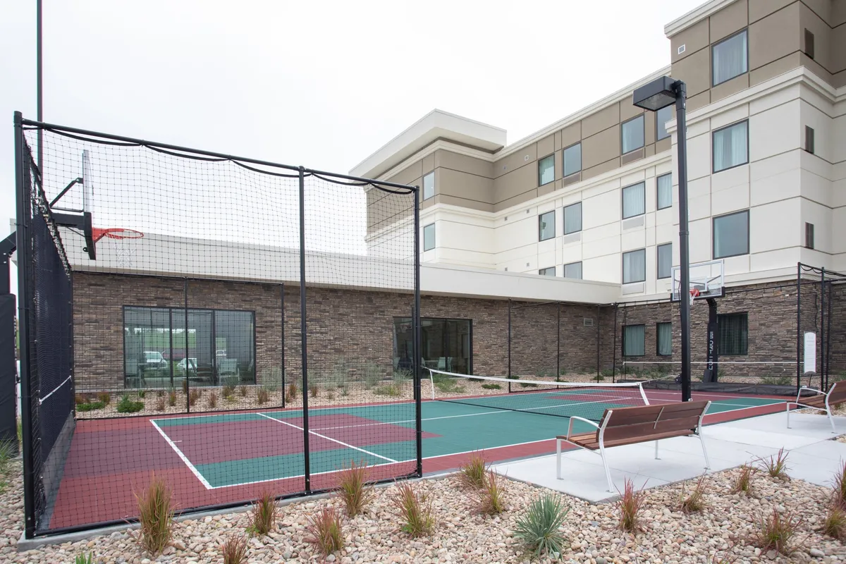 This vibrant exterior image captures the hotel's recreational sport court area, featuring a basketball hoop, tennis net, benches, and neatly landscaped surroundings. Guests are depicted socializing, participating in energetic activities, and enjoying open-air amenities.