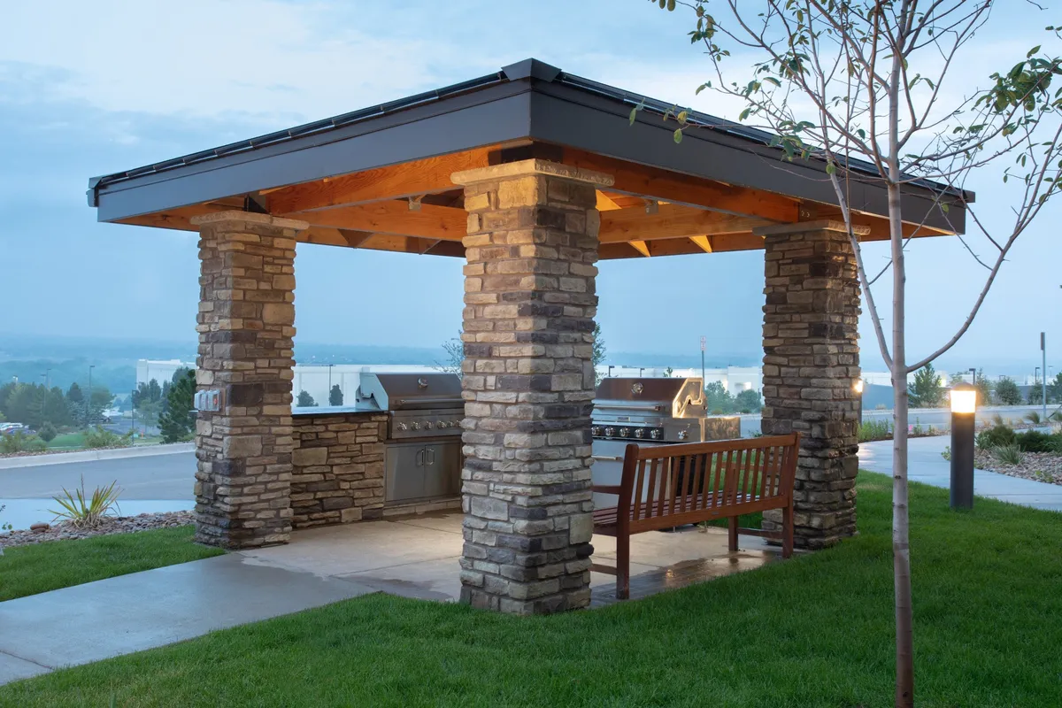 An inviting outdoor scene featuring sturdy stone pillars, a communal grill area, and a wooden bench set against a sunset backdrop. Guests are depicted enjoying a relaxed barbecue gathering, fostering an atmosphere of social connection and outdoor leisure.