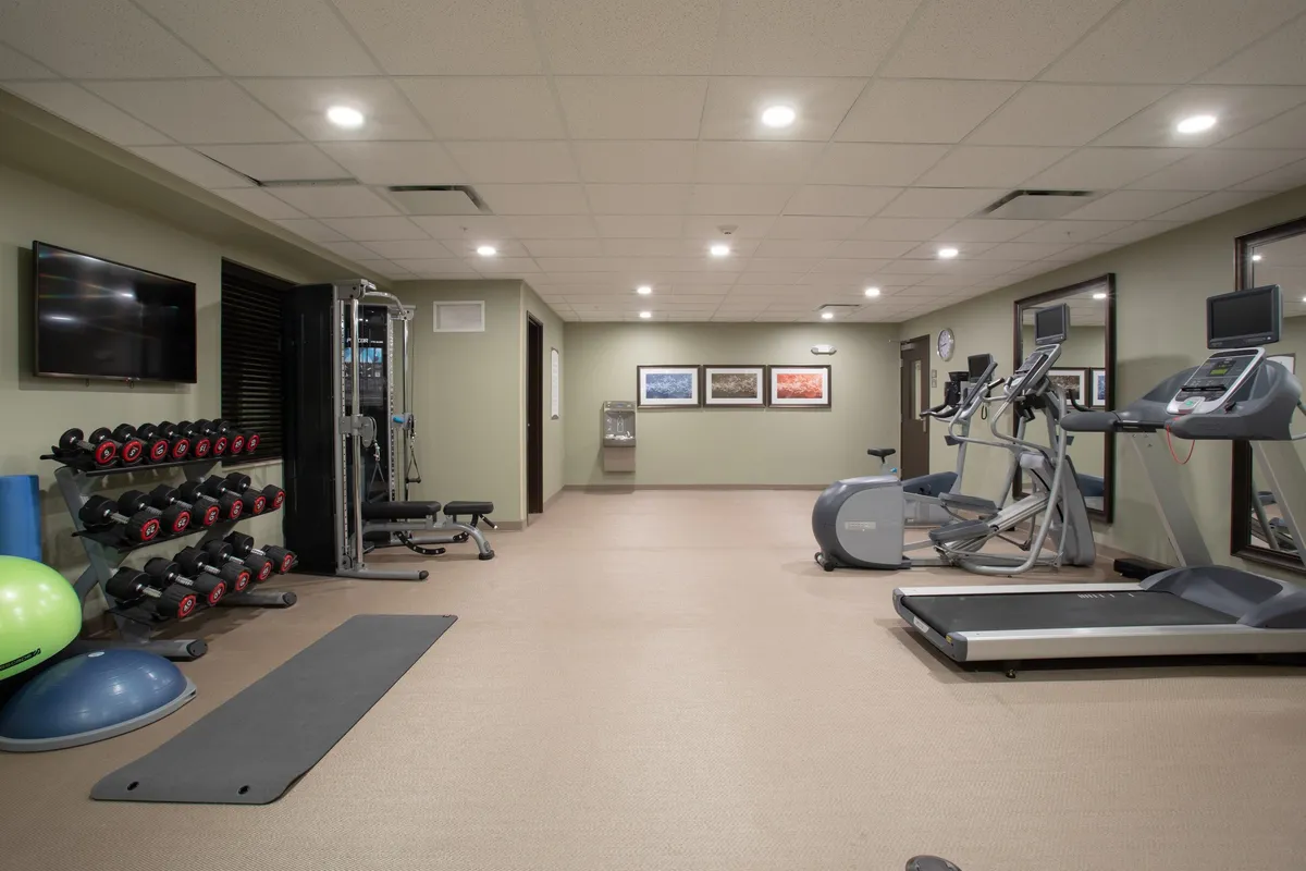 A well-equipped fitness center with a variety of workout equipment, including a dumbbell rack, treadmills, elliptical machines, exercise mats, a water cooler, and motivational wall art. The space is bright and encourages both individual and group exercise.