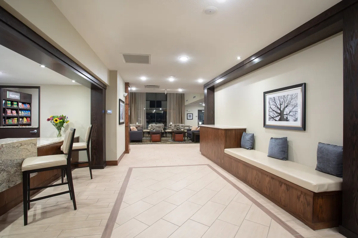 A welcoming reception area featuring a counter stocked with snacks, comfortable plush chairs, bench seating with colorful cushions, and modern artwork on the wall. The space is softly lit and designed for guests to relax, socialize, or grab a refreshment.